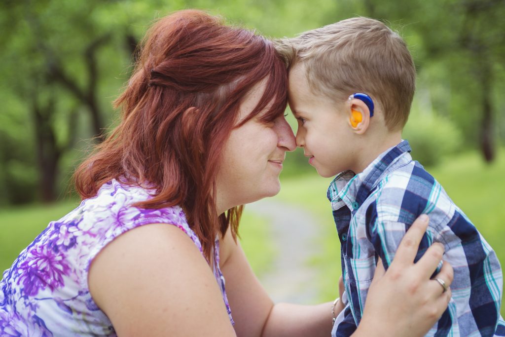 A Mother and young son with a hearing aid, in forest. They are pressing their forheads